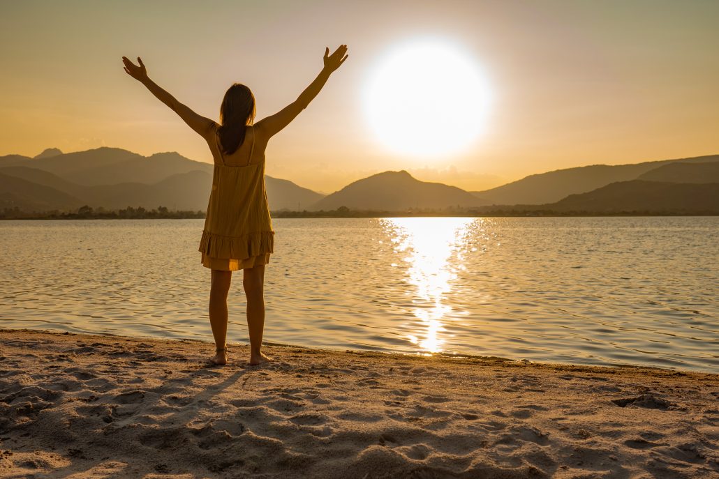 A person in a yellow dress stands by a lake at sunset, arms raised in a gesture of joy or celebration.