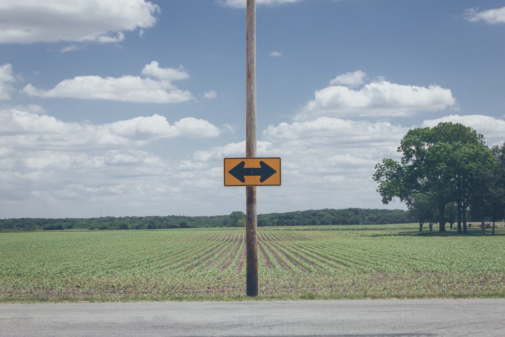 Roadside sign with arrows pointing left and right, surrounded by an open field under a blue sky.