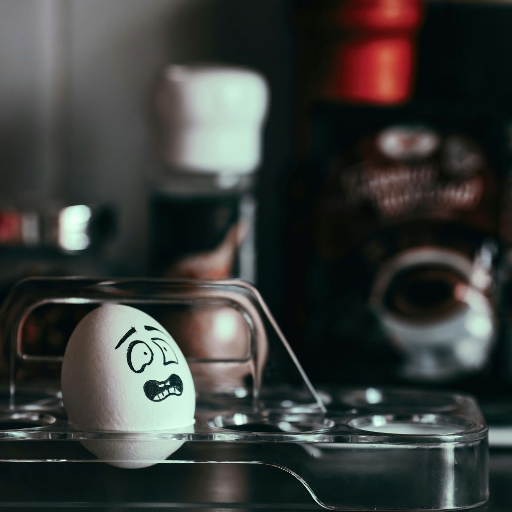 A cartoon-illustrated egg with a worried expression sits in an egg tray among jars and bottles in a kitchen setting.