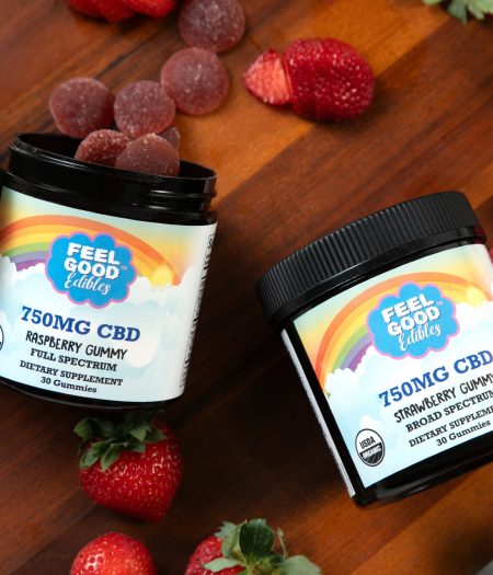 Raspberry and strawberry CBD gummy jars on a wooden surface, surrounded by fresh strawberries and a cup of coffee.